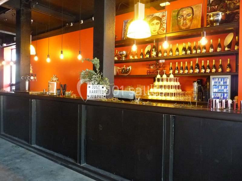 Bar avec comptoir noir, mur orange, étagères garnies de bouteilles, décorations et présentoir à coupes de champagne.