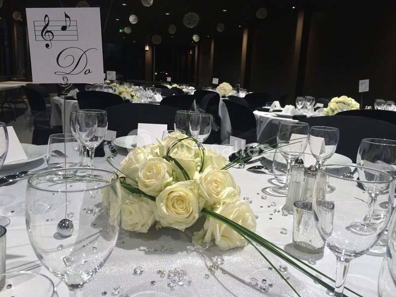 Salle de réception décorée avec des tables élégantes, ornées de roses blanches, perles et menus musicaux.