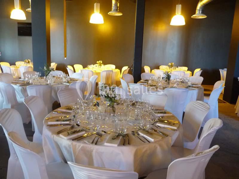 Salle de réception avec des tables rondes dressées, nappes blanches, chaises assorties et décorations florales.