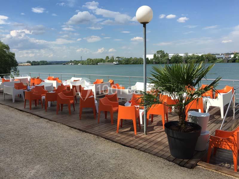 Terrasse en bois avec des chaises orange et blanches, vue sur une rivière bordée d'arbres sous un ciel dégagé.