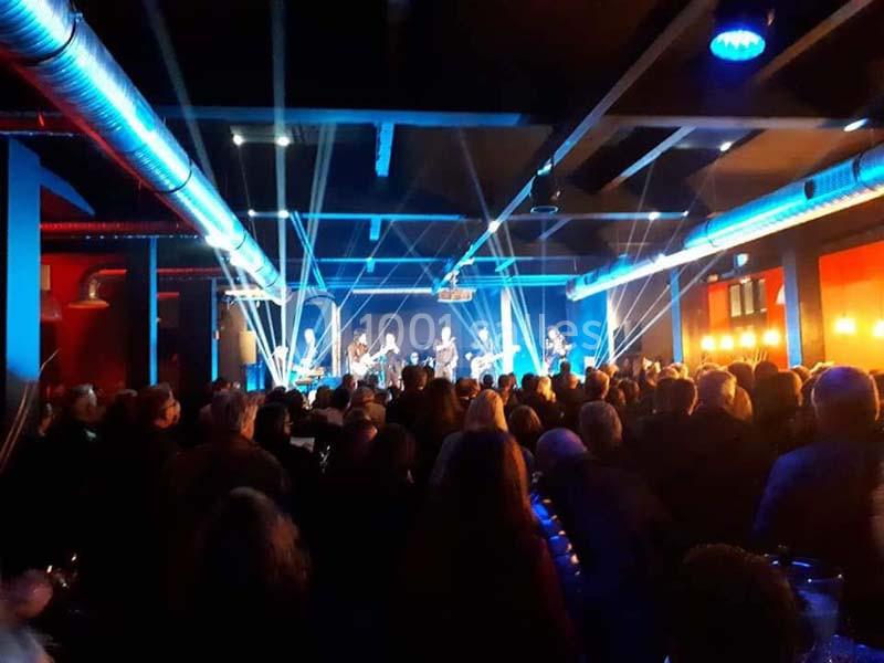 Salle de concert bondée avec un groupe jouant sur scène sous des éclairages colorés et des faisceaux lumineux.