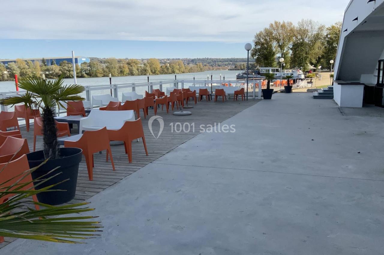 Terrasse avec chaises orange et tables blanches en bord de rivière, entourée de végétation et d'un ciel dégagé.