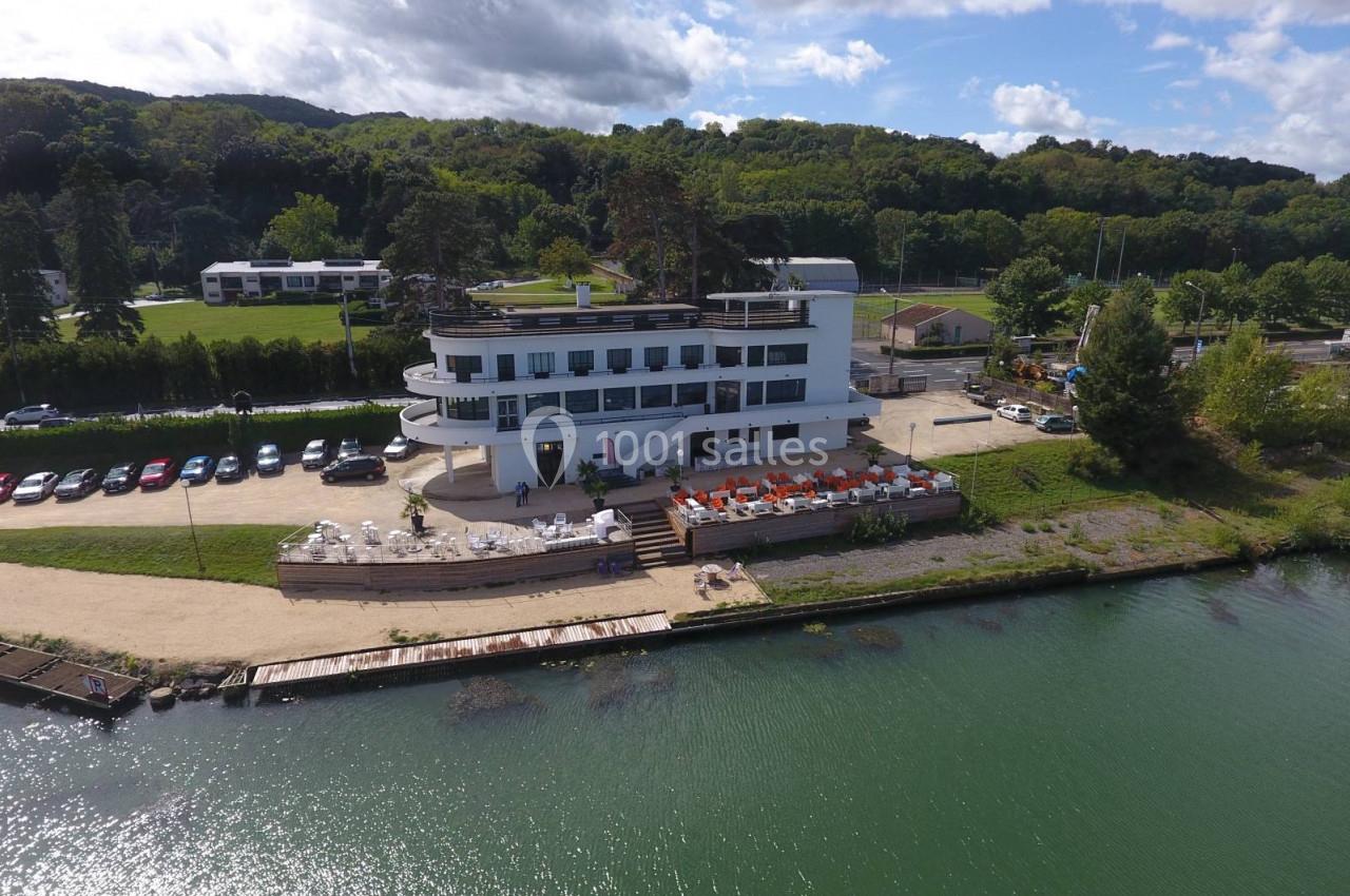 Bâtiment blanc moderne en bord de rivière, entouré de verdure, avec une terrasse et un parking adjacent.