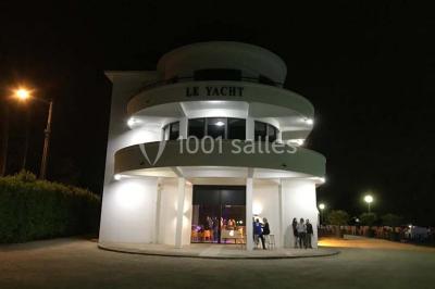 Façade éclairée d'un bâtiment nommé ’Le Yacht’ de nuit, avec des lumières bleues et un espace extérieur aménagé.