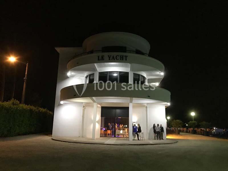 Bâtiment circulaire éclairé de nuit avec l'inscription ’Le Yacht’, des personnes rassemblées près de l'entrée.