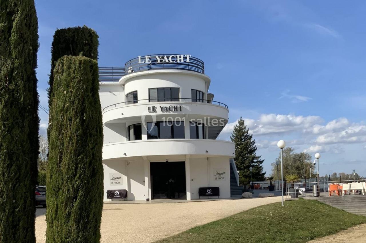 Bâtiment blanc moderne avec l'inscription ’Le Yacht’, entouré de cyprès et situé dans un espace dégagé sous un ciel bleu.