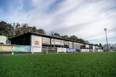 Deux hommes en tenue de rugby bleue simulent une action de jeu dans une salle éclairée avec des chaises vides.
