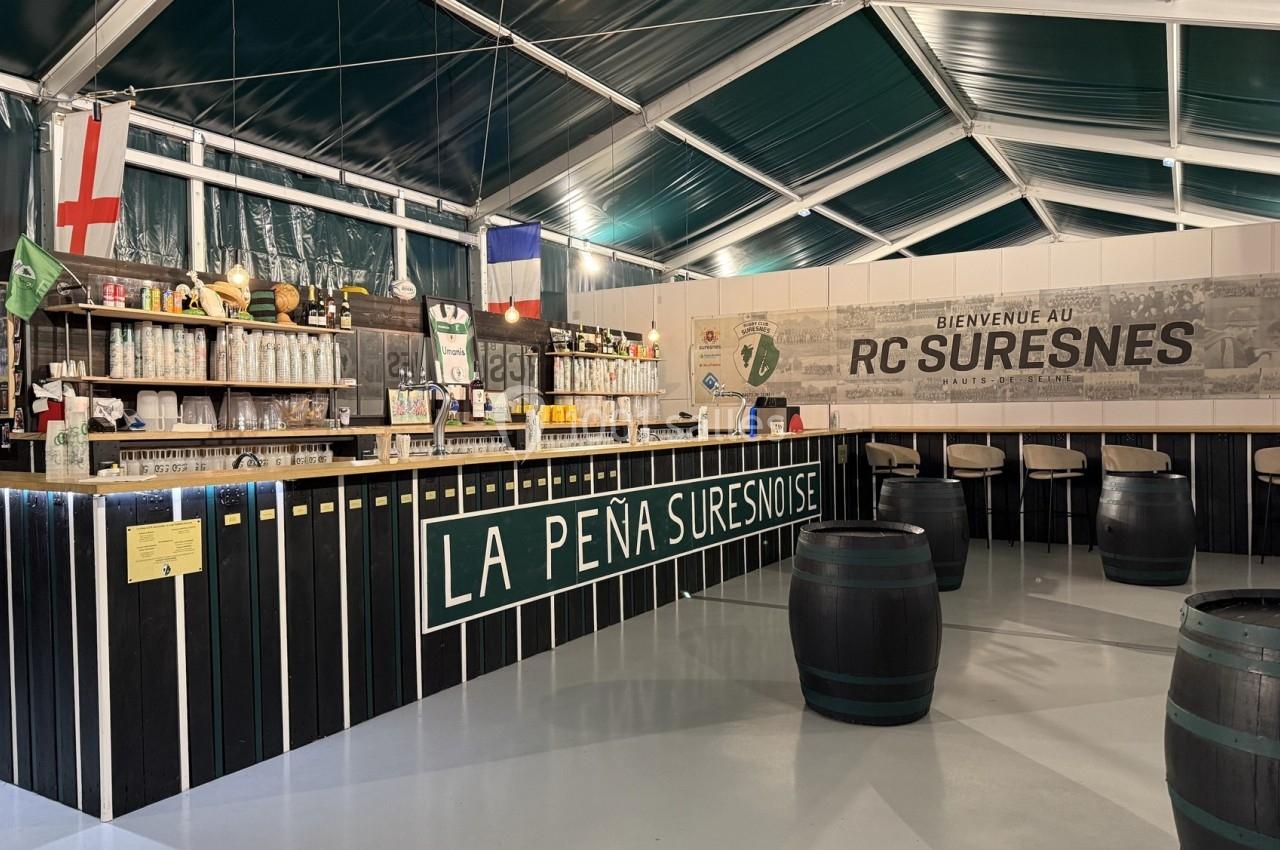 Intérieur d'un bar associatif avec comptoir en bois, tonneaux en guise de tables et décorations aux couleurs locales.