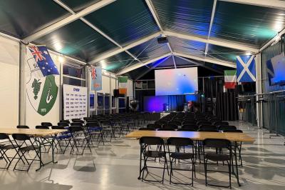Deux hommes en tenue de rugby bleue simulent une action de jeu dans une salle éclairée avec des chaises vides.