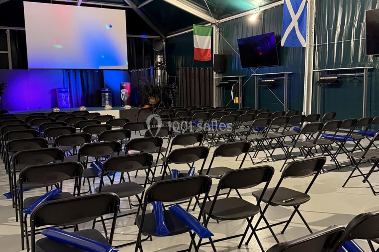 Salle de conférence vide avec des rangées de chaises noires, un écran géant et des drapeaux italien et écossais accrochés.