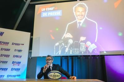 Deux hommes en tenue de rugby bleue simulent une action de jeu dans une salle éclairée avec des chaises vides.