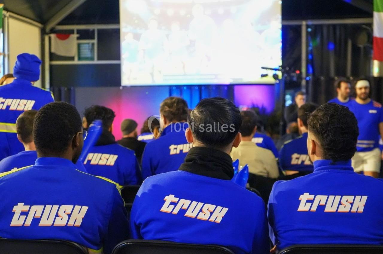 Des personnes en tenue bleue avec le logo ’Trusk’ regardent un écran géant dans une salle éclairée.
