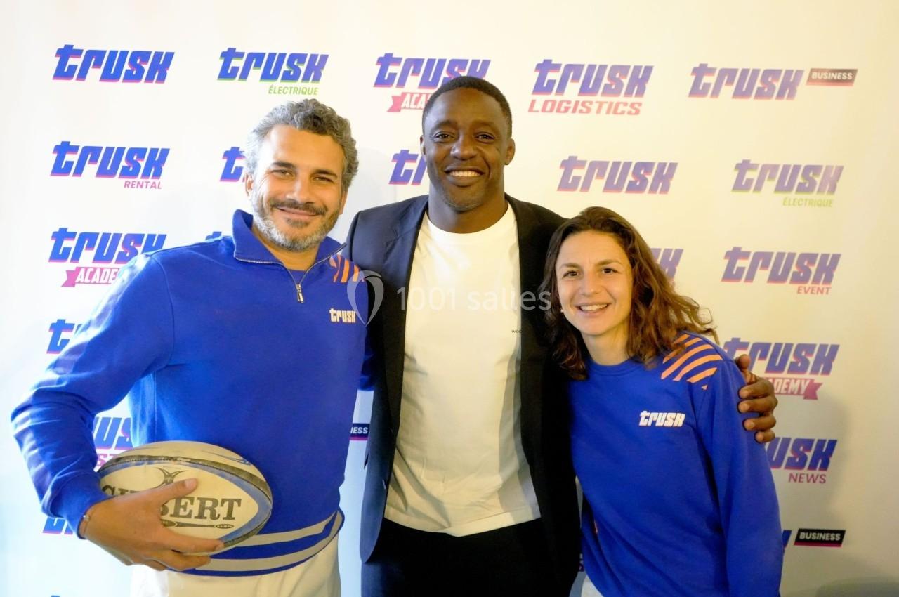 Trois personnes souriantes posent devant un fond avec des logos ’Trusk’, dont une tenant un ballon de rugby.