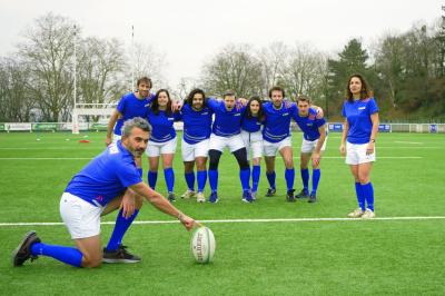 Deux hommes en tenue de rugby bleue simulent une action de jeu dans une salle éclairée avec des chaises vides.