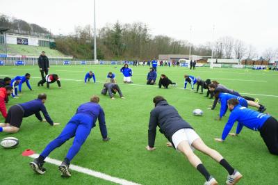 Deux hommes en tenue de rugby bleue simulent une action de jeu dans une salle éclairée avec des chaises vides.