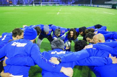 Deux hommes en tenue de rugby bleue simulent une action de jeu dans une salle éclairée avec des chaises vides.