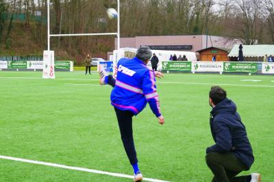 Deux hommes en tenue de rugby bleue simulent une action de jeu dans une salle éclairée avec des chaises vides.