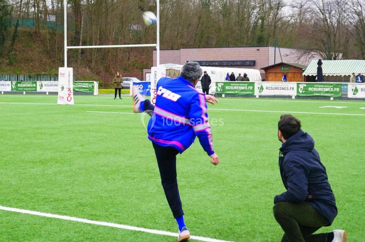 Un homme en veste bleue frappe un ballon de rugby sur un terrain, sous le regard d'une autre personne accroupie.