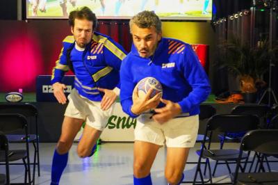 Deux hommes en tenue de rugby bleue simulent une action de jeu dans une salle éclairée avec des chaises vides.