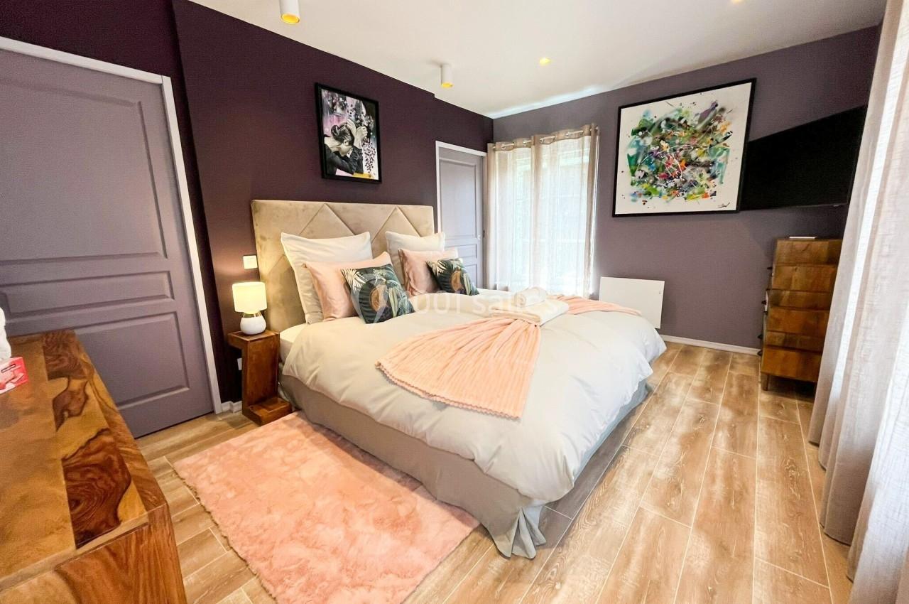 Chambre avec lit double, murs violets, parquet clair, tableaux décoratifs et lumière naturelle entrant par une fenêtre.