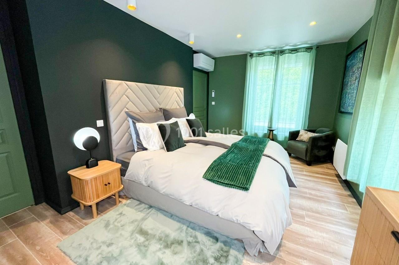 Chambre moderne avec lit double, tête de lit capitonnée, murs verts, parquet clair et grande fenêtre avec rideaux.