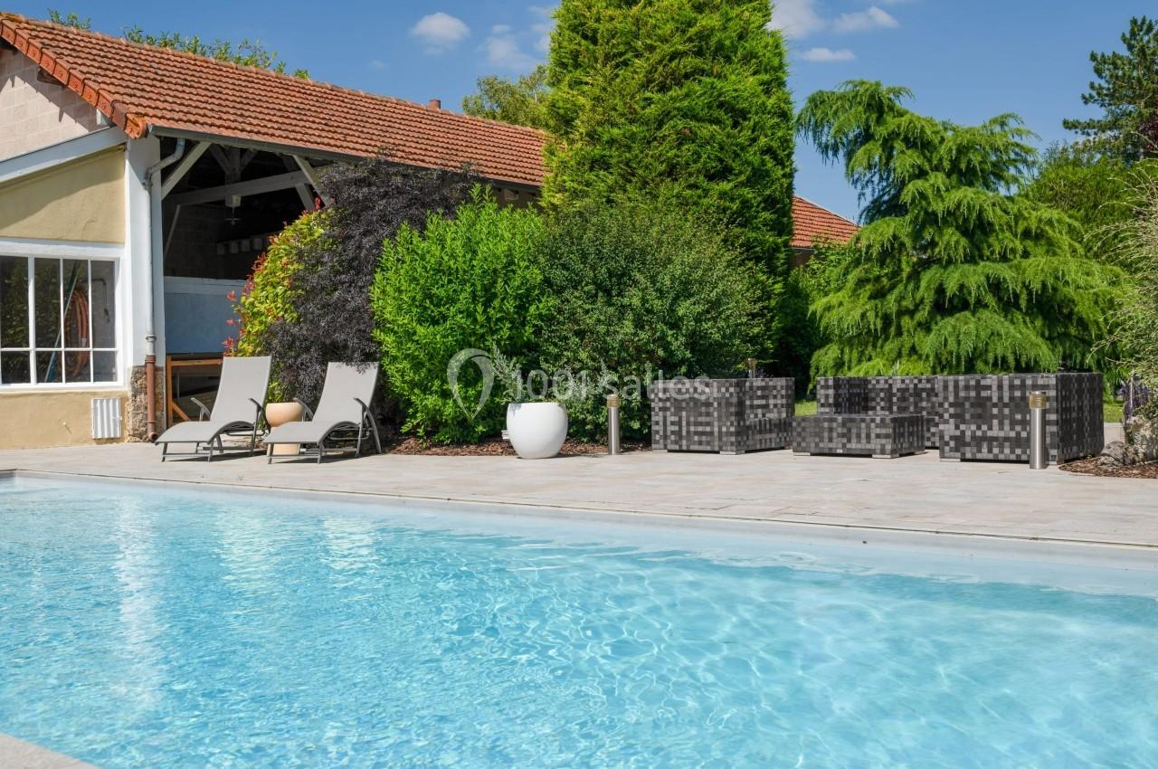 Piscine extérieure avec terrasse, chaises longues, mobilier de jardin et végétation luxuriante en arrière-plan.