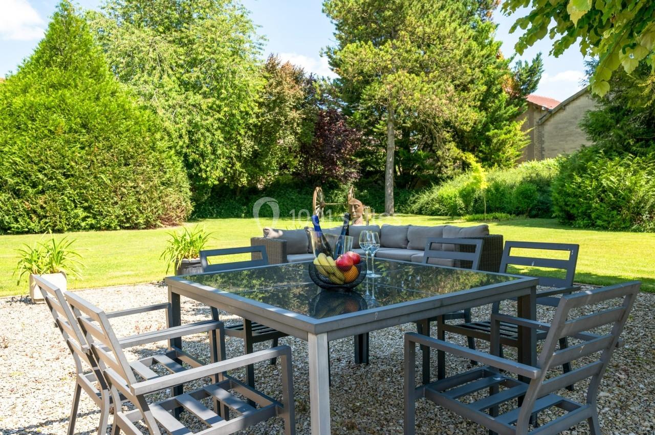 Table de jardin en métal avec chaises, décorée d'un panier de fruits, sur une terrasse en gravier entourée de verdure.