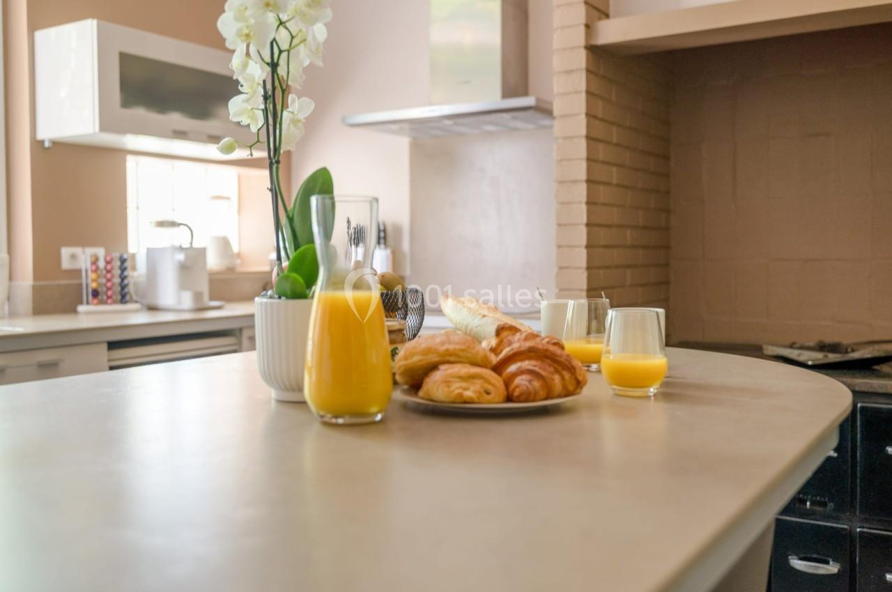 Petit-déjeuner avec croissants, baguette, jus d'orange et orchidée sur un plan de travail dans une cuisine lumineuse.