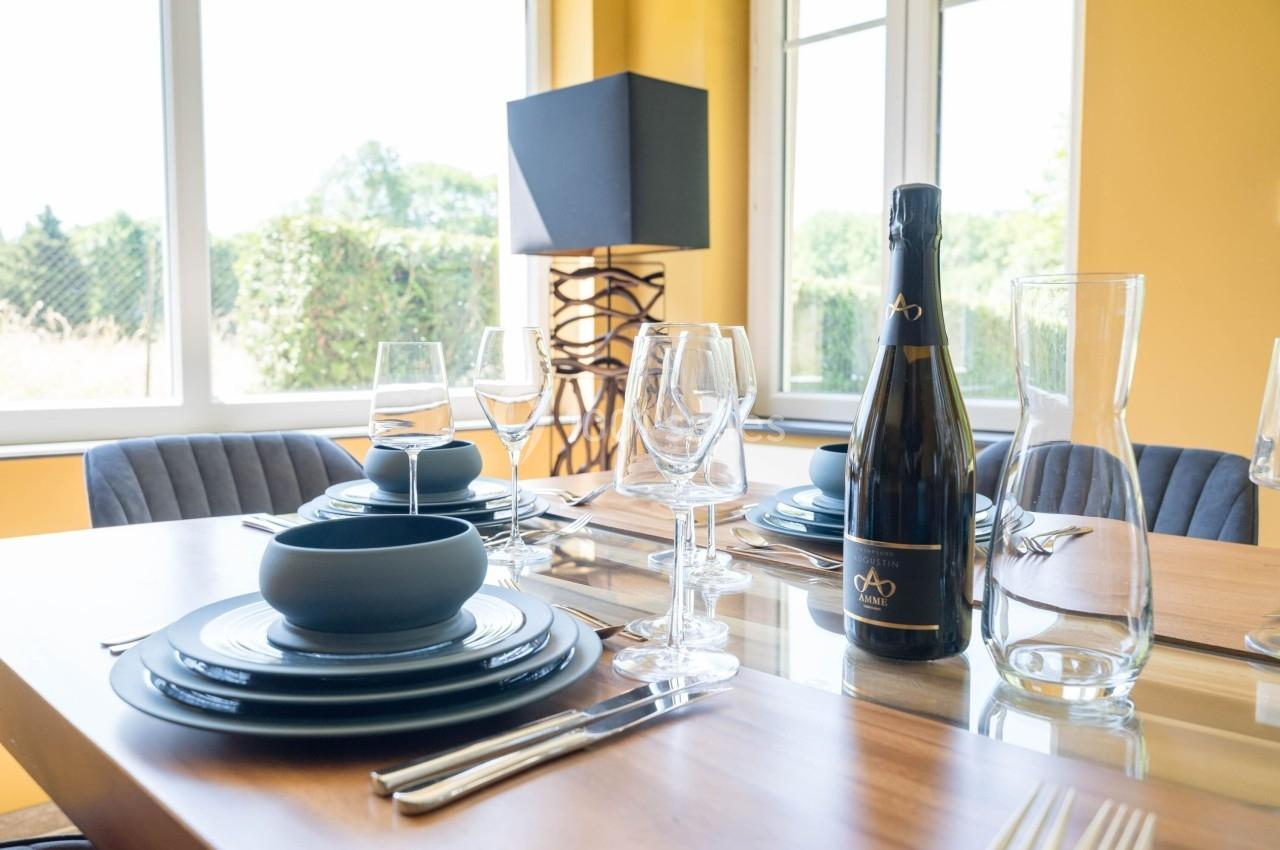 Table dressée avec vaisselle noire, verres à pied, carafe et bouteille de vin dans une salle lumineuse aux murs jaunes.