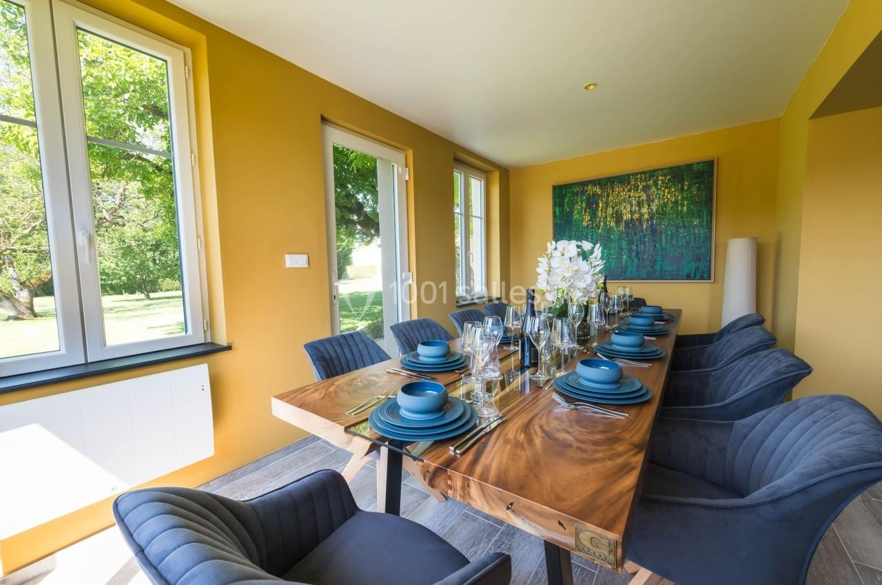 Salle à manger lumineuse avec murs jaunes, table en bois dressée de vaisselle bleue et chaises rembourrées grises.