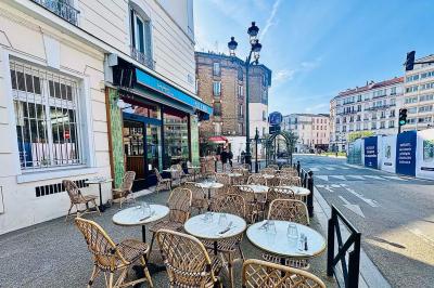 Terrasse de café avec tables et chaises en osier, située sur un trottoir ensoleillé dans un quartier urbain.