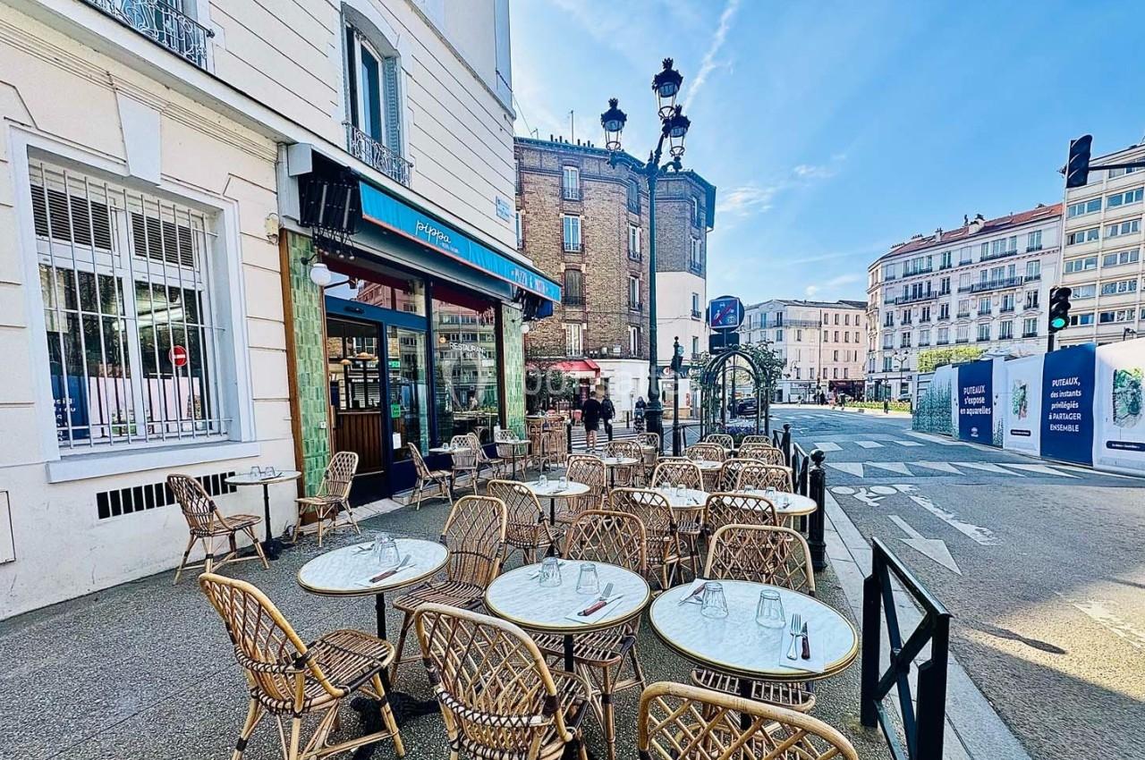 Terrasse de café avec tables et chaises en osier, située sur un trottoir ensoleillé dans un quartier urbain.