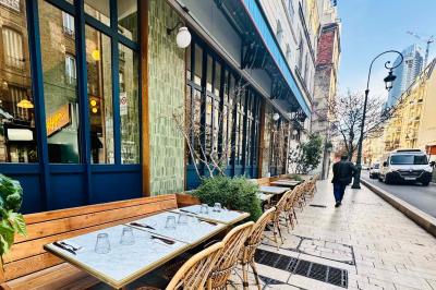 Terrasse de café avec tables et chaises en osier, située sur un trottoir ensoleillé dans un quartier urbain.