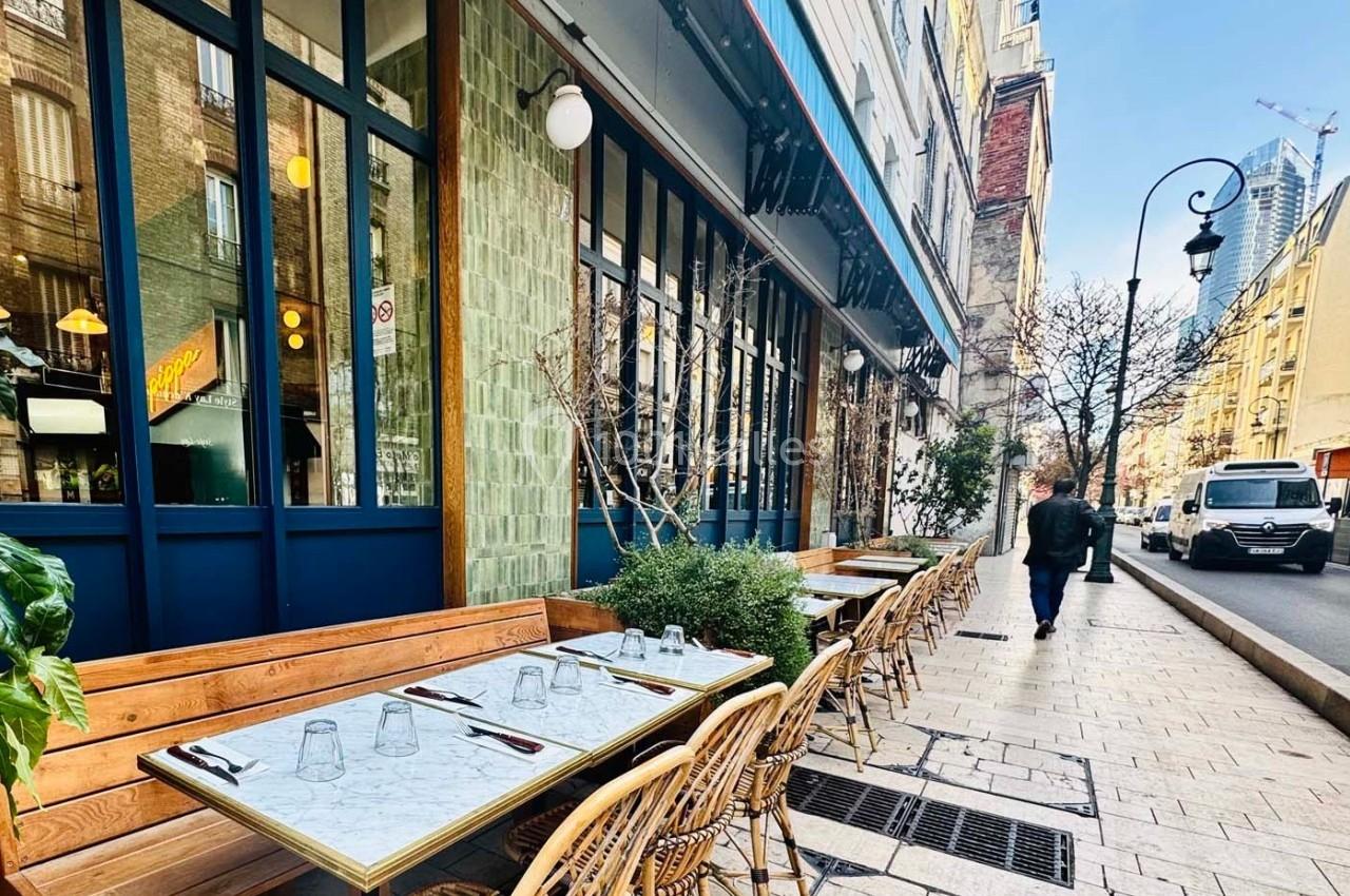 Terrasse de restaurant avec tables dressées, située dans une rue pavée bordée de bâtiments et d'arbres.