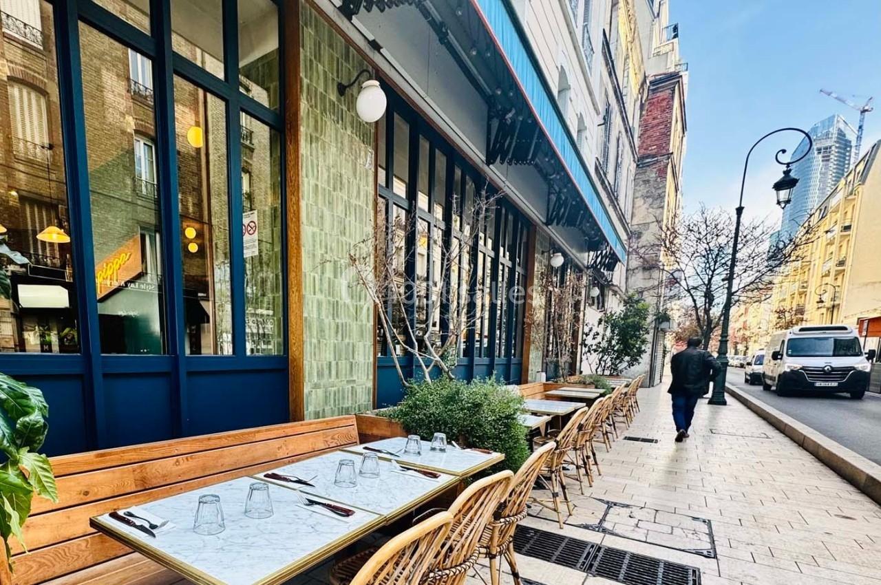 Terrasse de café avec tables en marbre et chaises en osier, située le long d'une rue pavée bordée de bâtiments urbains.