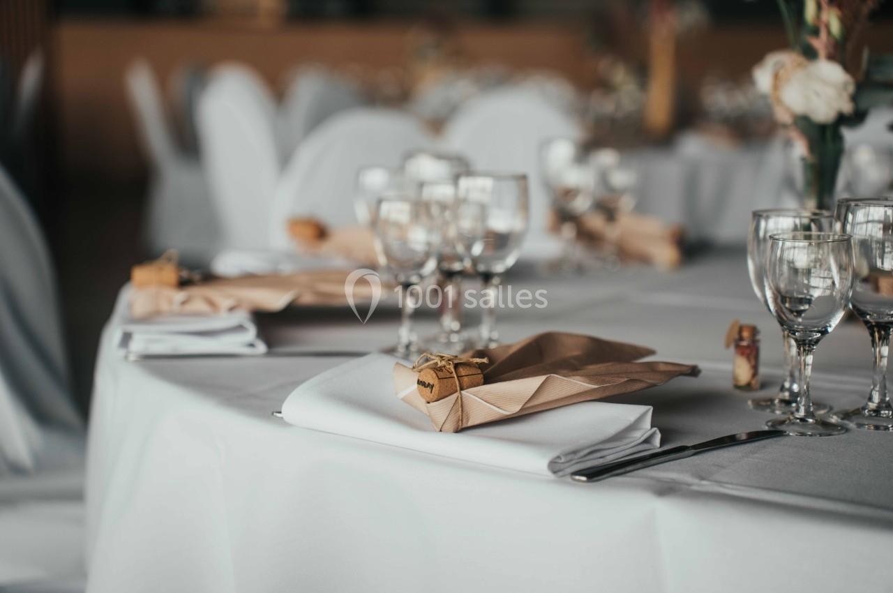 Table dressée avec nappes blanches, serviettes pliées, verres à pied et décorations élégantes dans une salle de réception.