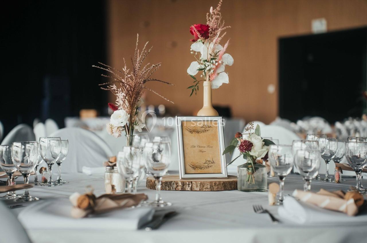 Table décorée pour un événement avec fleurs, verres, serviettes pliées et un cadre posé sur une rondelle de bois.