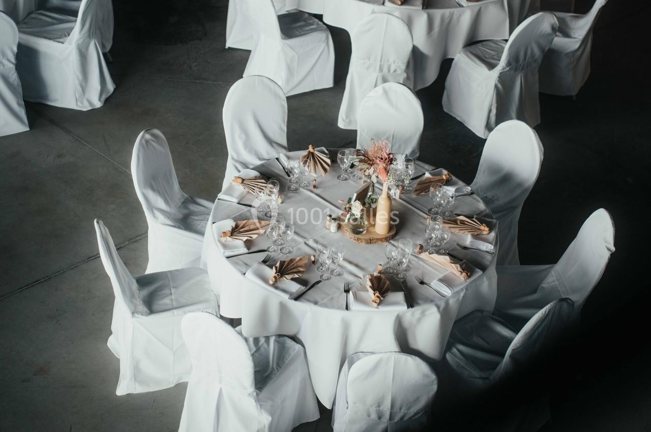Table ronde décorée avec nappes blanches, serviettes dorées et centre de table floral, entourée de chaises houssées.
