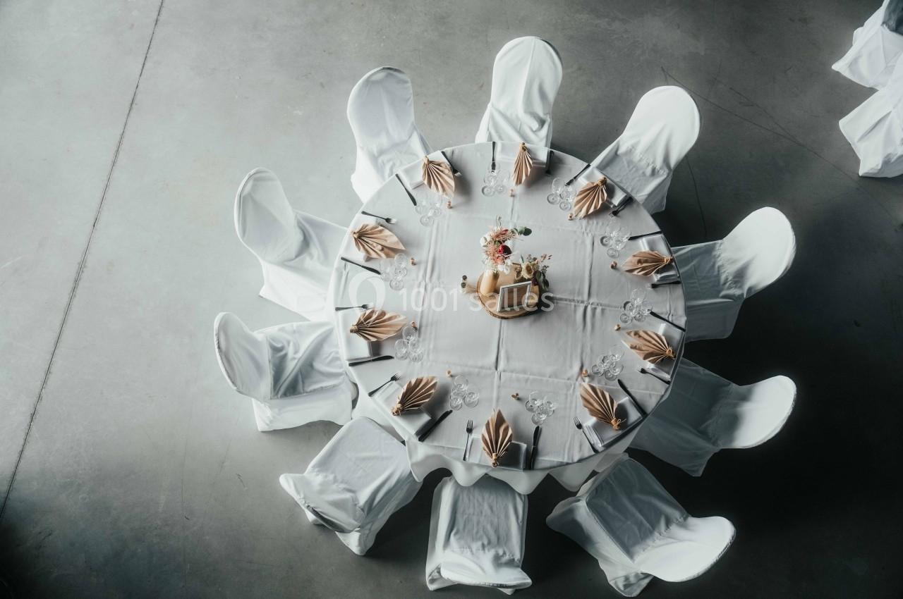 Table ronde dressée avec une nappe blanche, chaises couvertes, serviettes pliées et un centre de table floral.