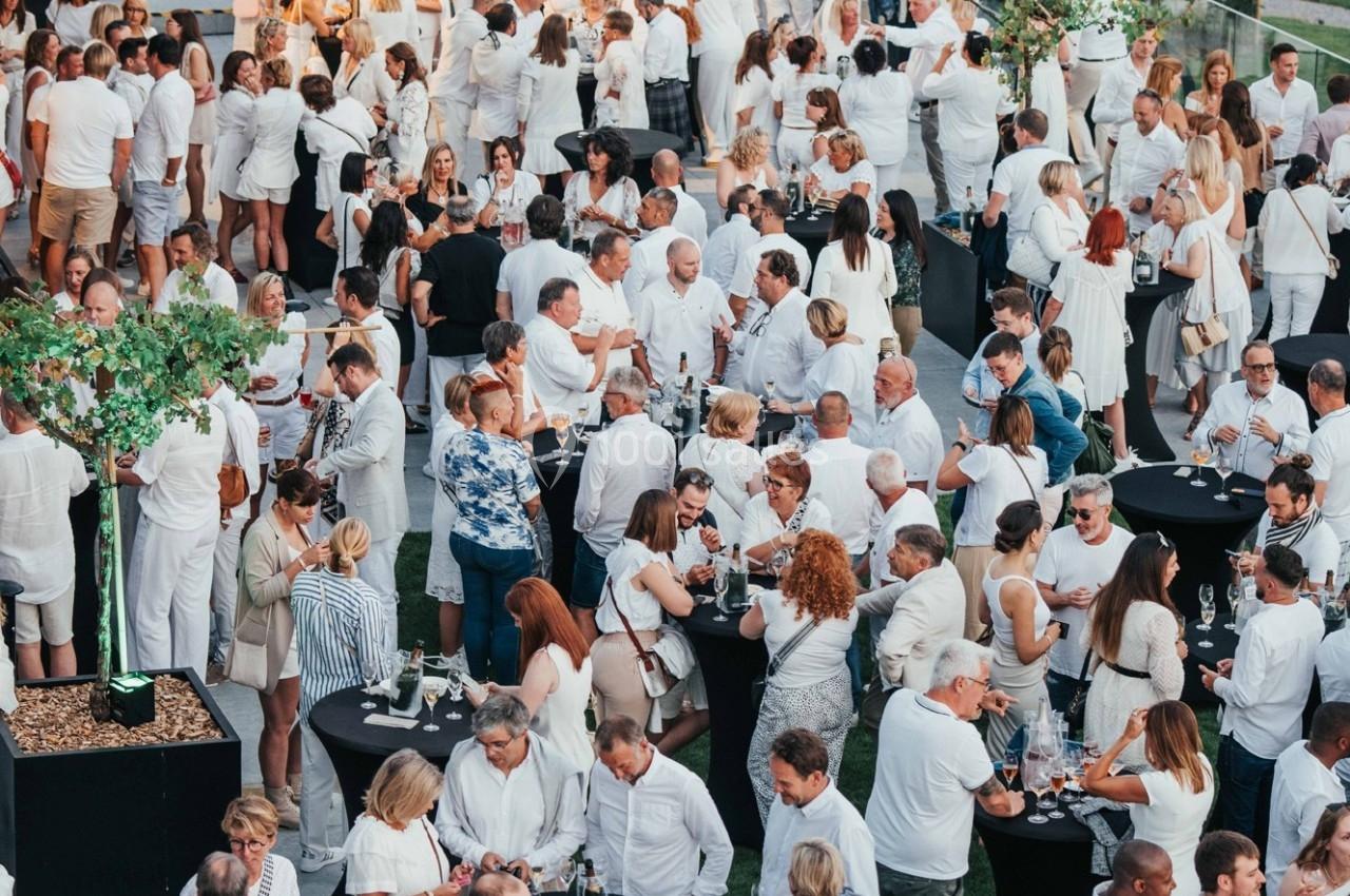 Groupe de personnes vêtues de blanc réunies lors d'un événement en plein air avec des tables hautes et des boissons.