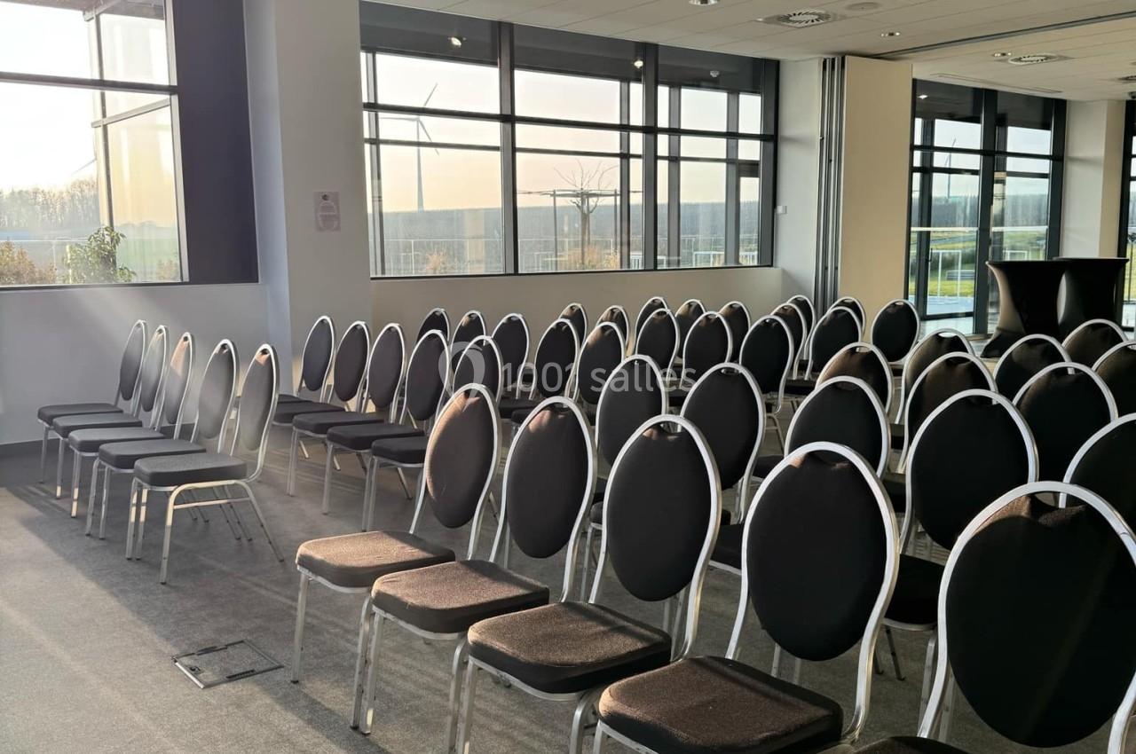 Salle de conférence vide avec des rangées de chaises noires alignées, éclairée par la lumière naturelle d'une grande fenêtre.