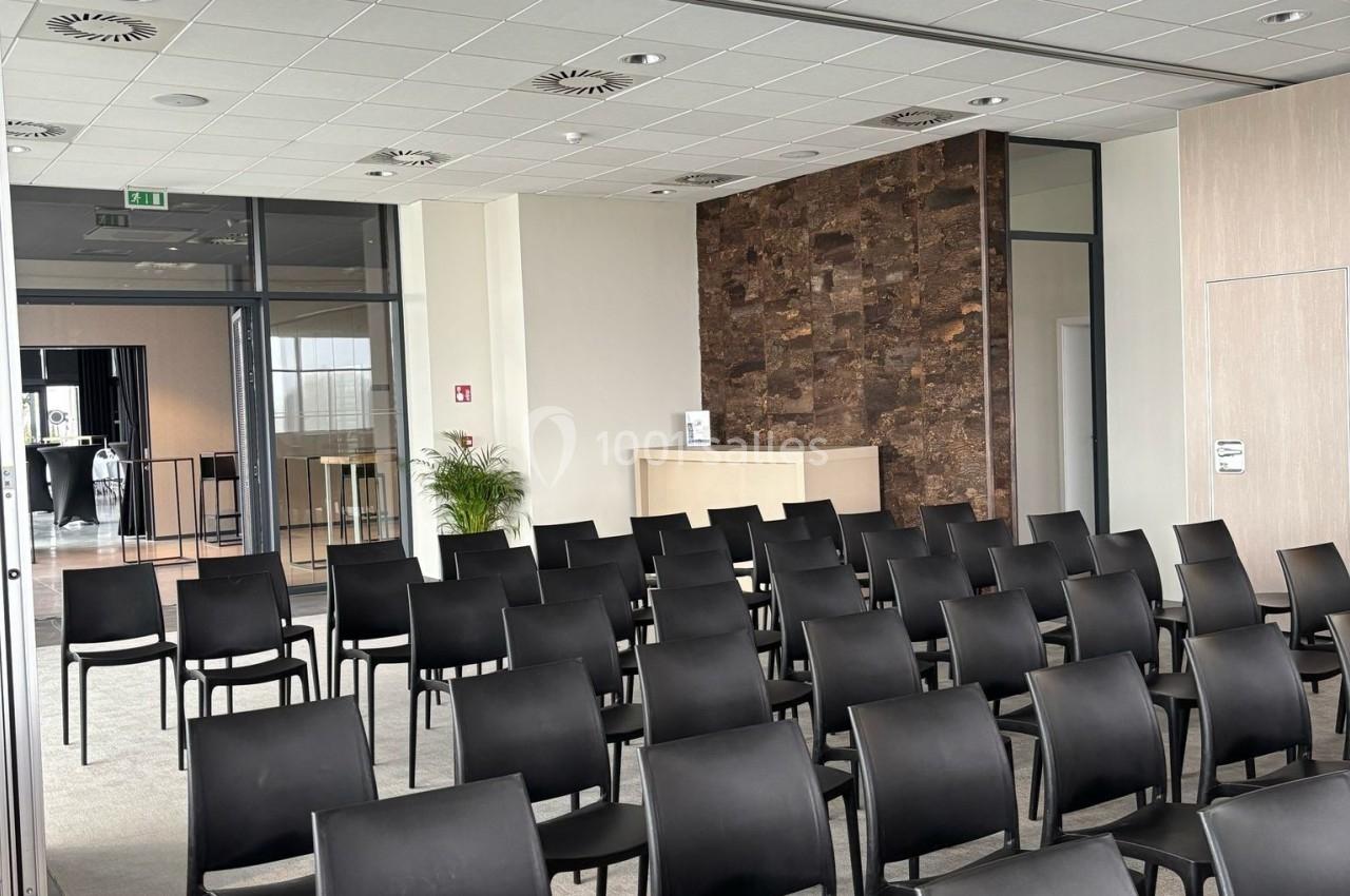 Salle de conférence moderne avec rangées de chaises noires alignées, murs clairs et comptoir en arrière-plan.
