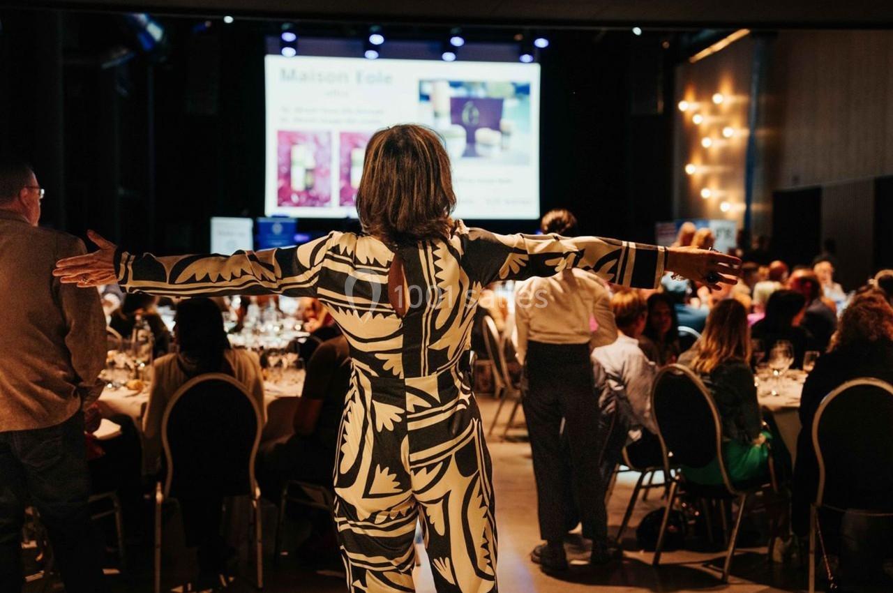 Une personne debout, bras écartés, face à un public assis dans une salle de conférence éclairée.