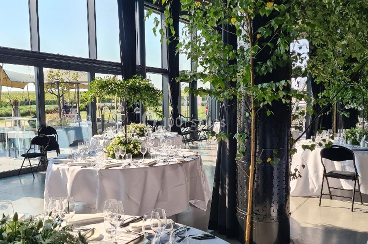 Salle lumineuse avec de grandes baies vitrées, tables élégamment dressées et décorations végétales.