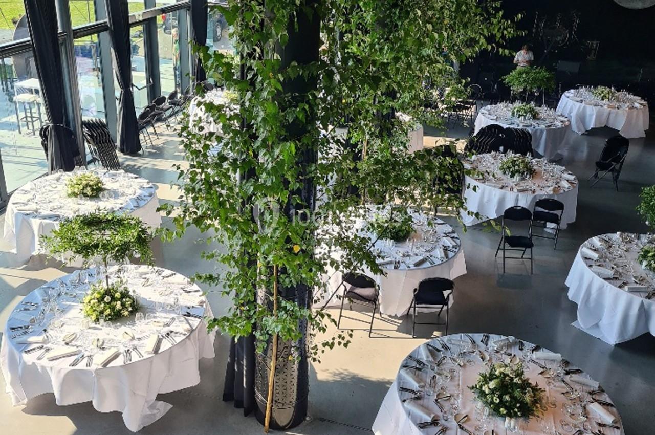 Salle de réception avec des tables rondes dressées, décorées de nappes blanches et de centres de table végétaux.