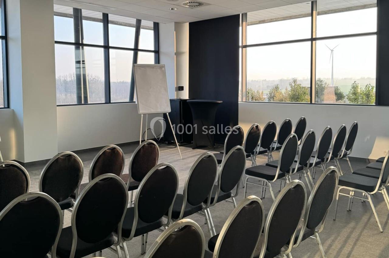 Salle de réunion lumineuse avec des rangées de chaises noires, un tableau blanc et une vue sur l'extérieur avec éolienne.
