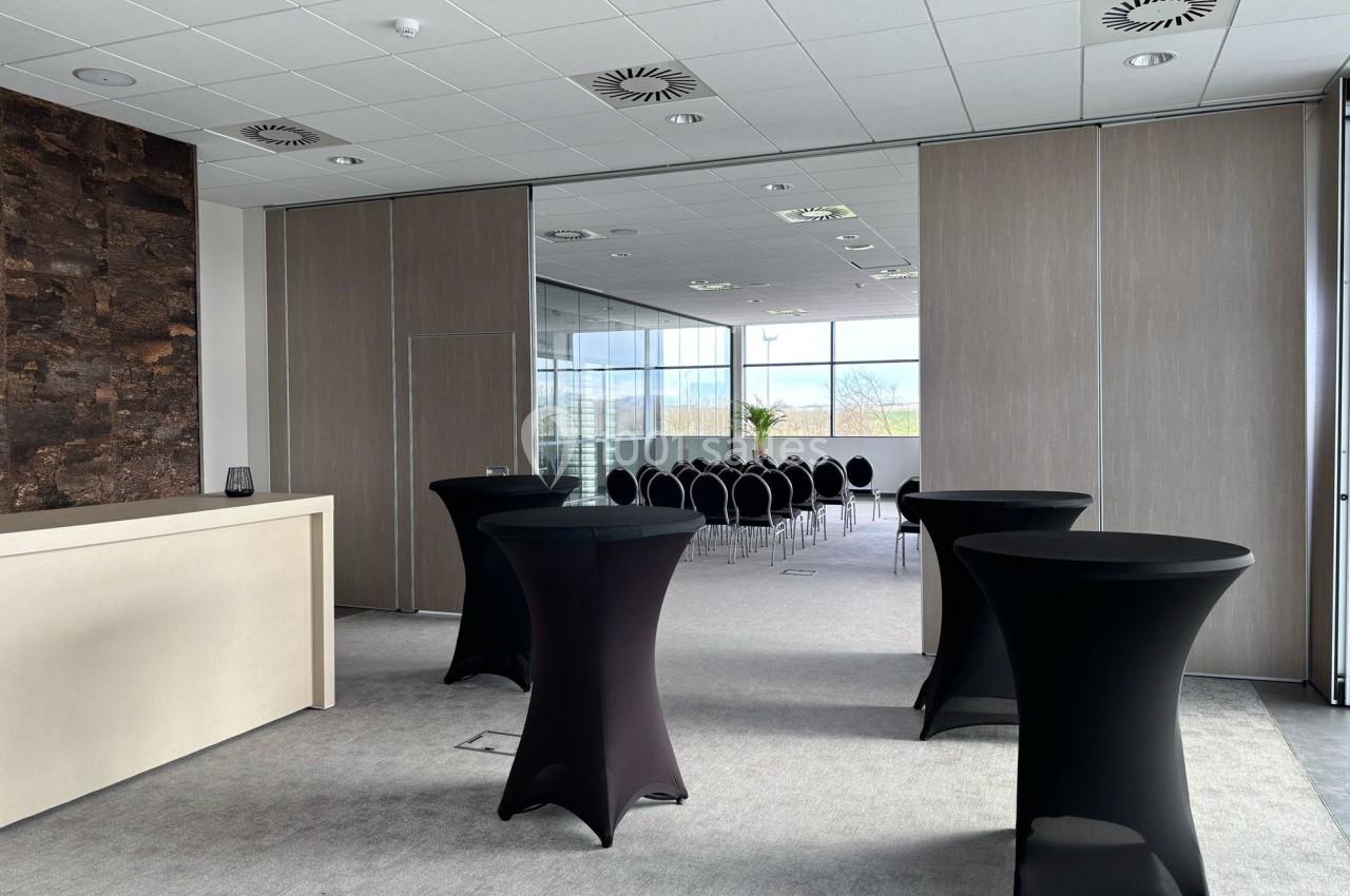 Salle de conférence lumineuse avec tables hautes noires et rangée de chaises alignées devant une baie vitrée.