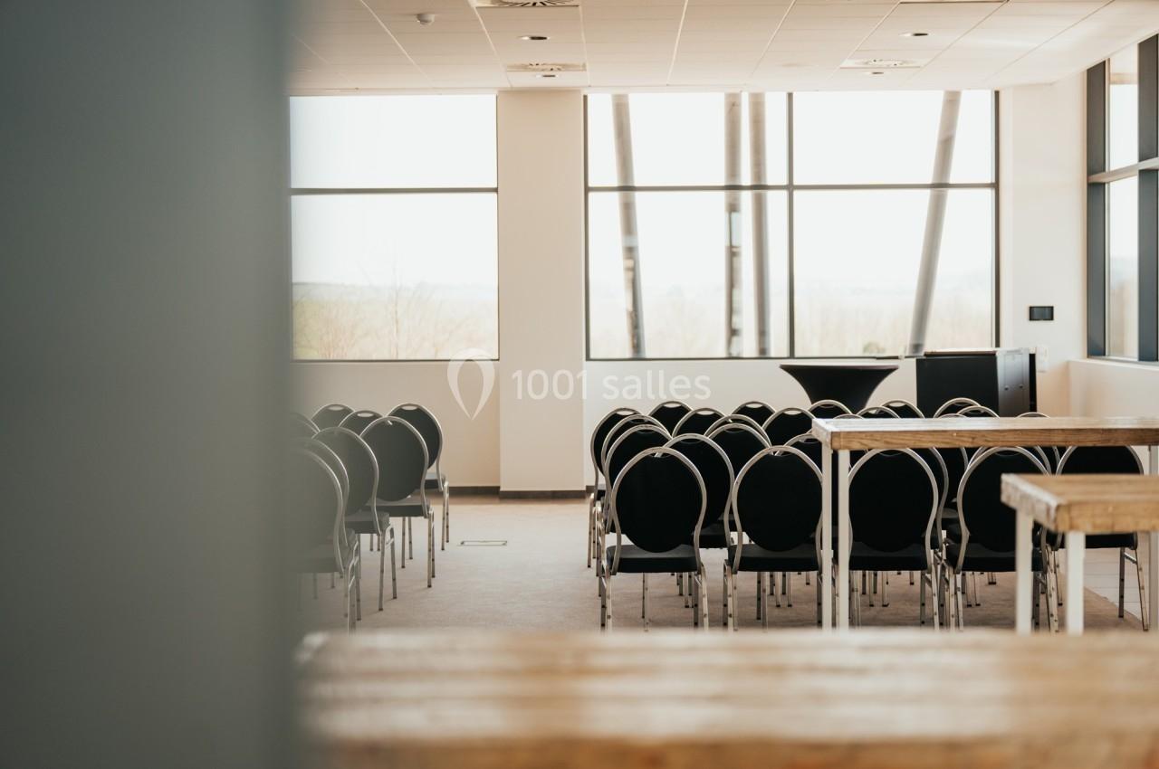Salle de conférence lumineuse avec des rangées de chaises noires alignées et de grandes fenêtres en arrière-plan.