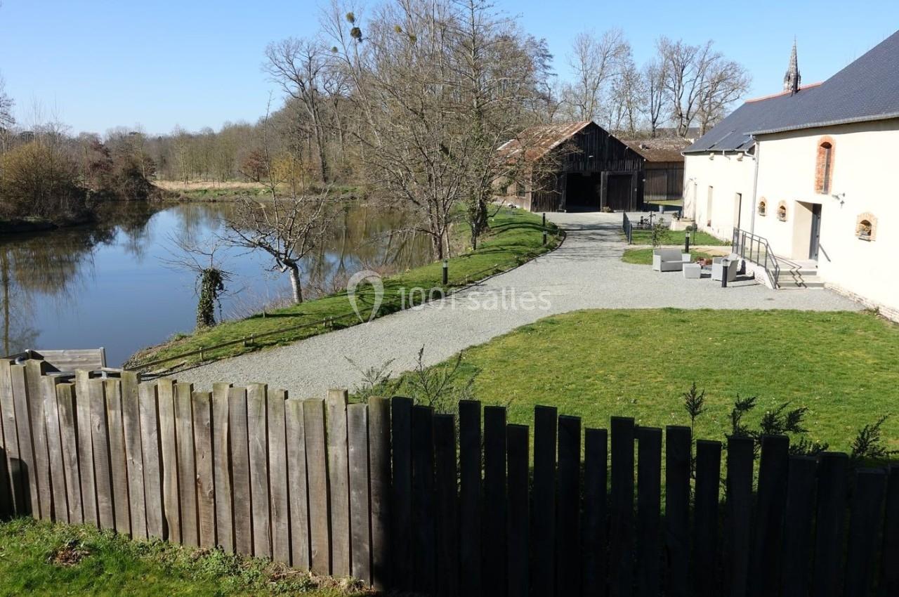 Vue d'un étang bordé d'arbres, avec une maison en pierre et une grange en arrière-plan, sous un ciel dégagé.