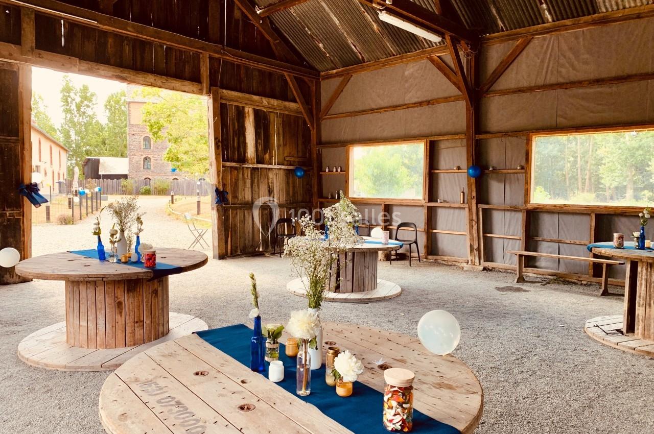 Intérieur d'une grange aménagée avec des tables rondes décorées de fleurs et de bocaux, éclairée par la lumière naturelle.